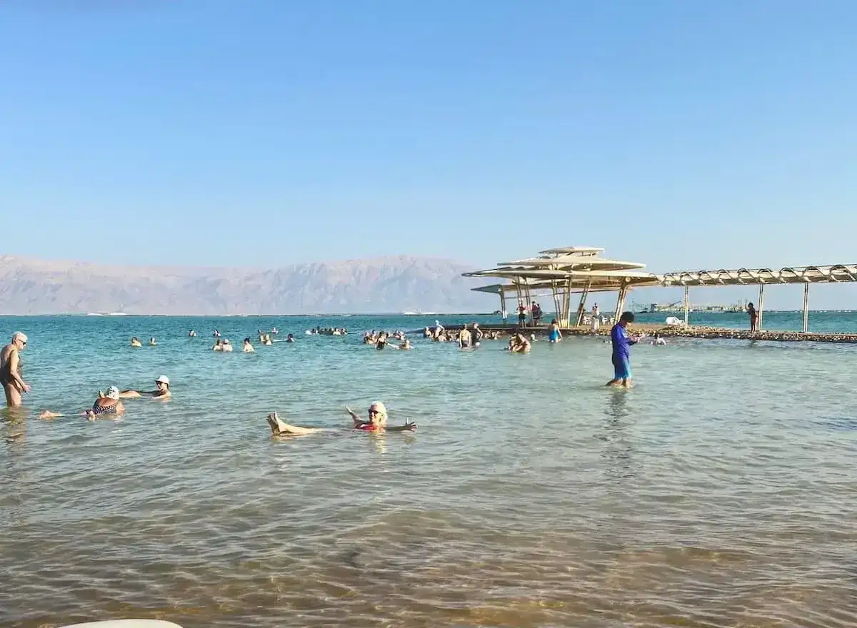 Ludzie unoszą się na wodzie Morza Martwego, korzystając z jego niezwykłego zasolenia. W tle góry i plaża z zacienionymi miejscami.