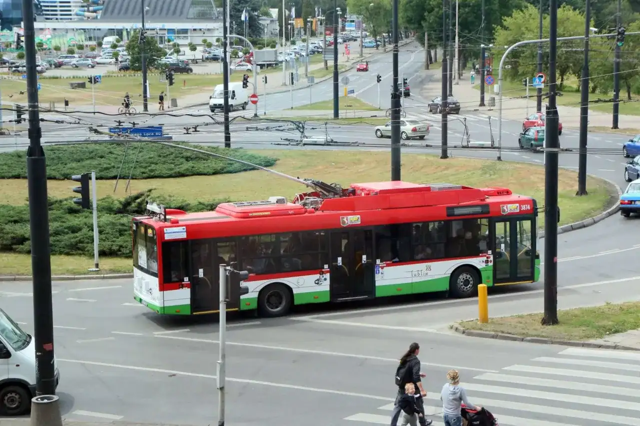 Czerwony trolejbus z napisem "Lublin" przejeżdża przez skrzyżowanie. W tle widać ruch uliczny i budynki.