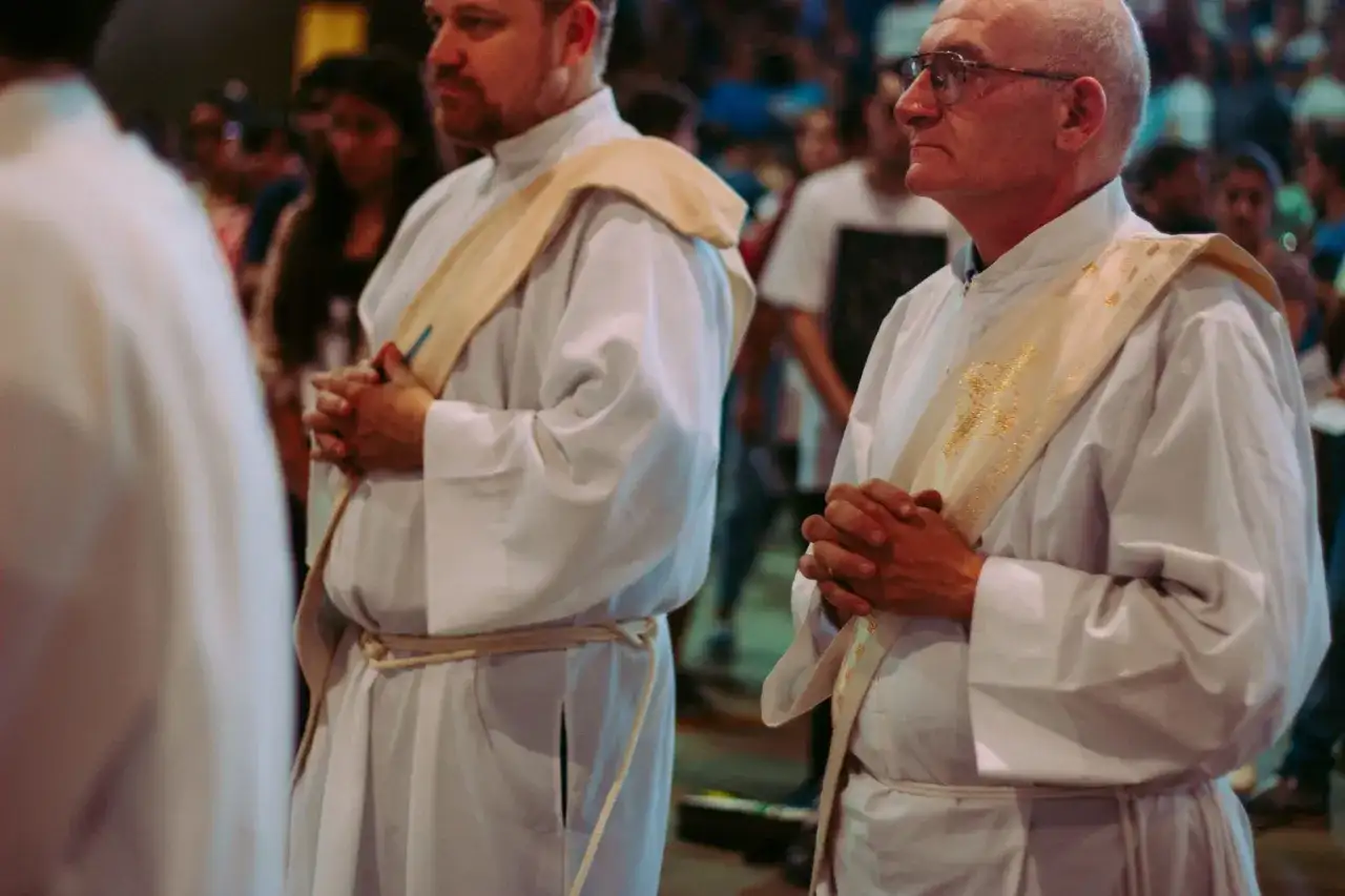 Diakoni i księża w białych szatach, z modlitewnie złożonymi dłońmi. Różnica między diakonem stałym a księdzem, ich obowiązki i przywileje.