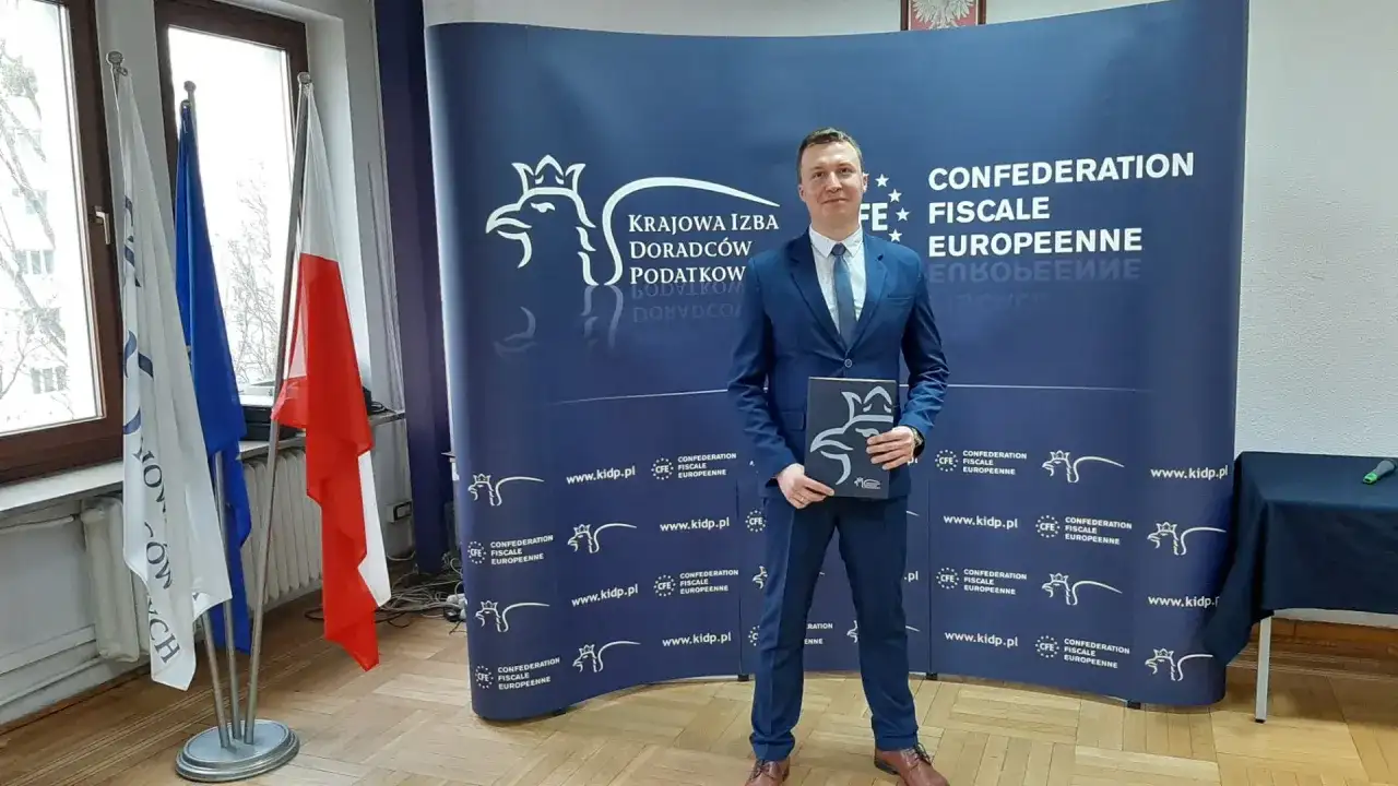 Mężczyzna w garniturze stoi przed niebieskim banerem z logo Krajowej Izby Doradców Podatkowych.