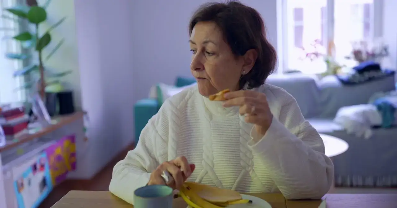 Kobieta w białym swetrze je banana, czując uczucie guli w gardle.