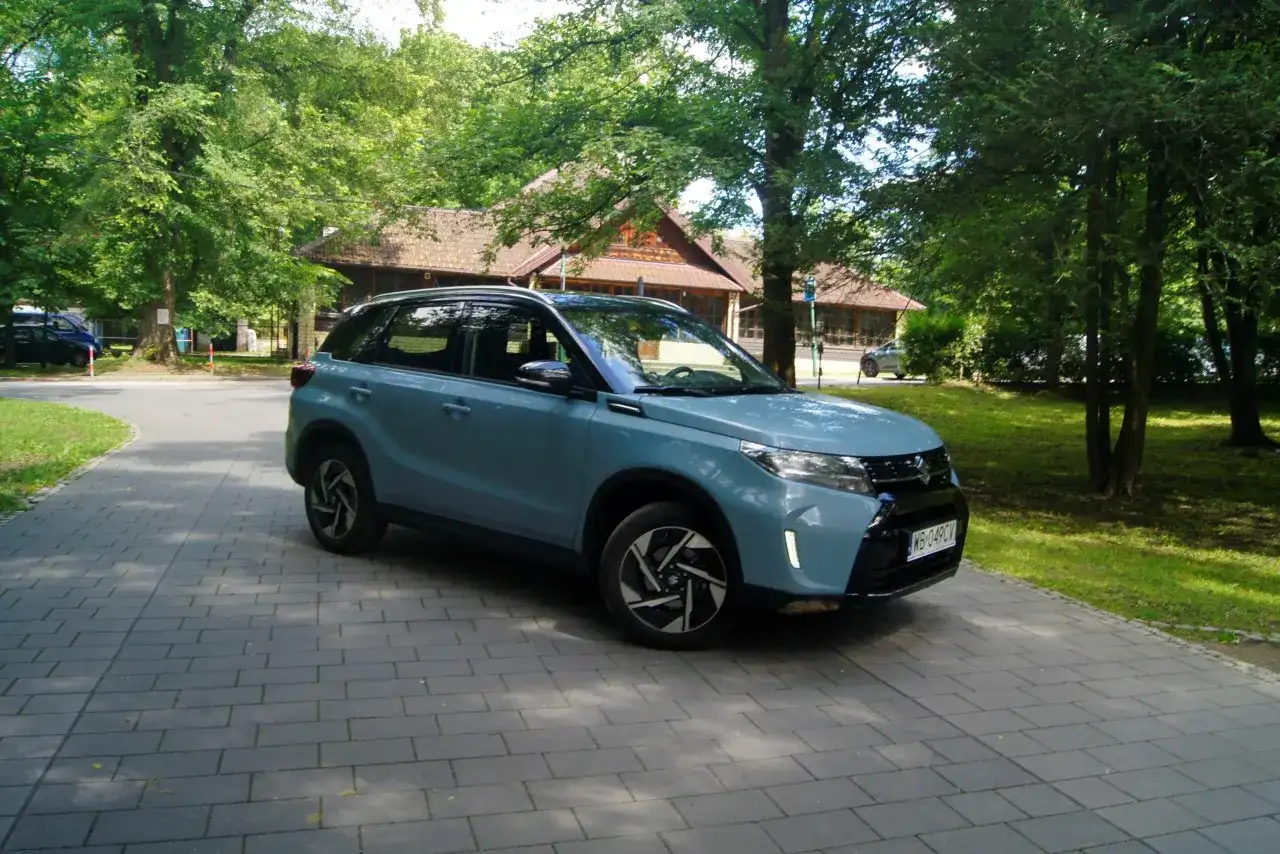 Jasnoniebieski Suzuki Vitara Hybrid zaparkowany na wybrukowanym podjeździe, otoczony zielenią i drewnianym budynkiem.
