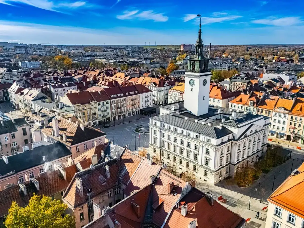 Widok z lotu ptaka na rynek z ratuszem i wieżą zegarową, otoczony kamienicami.
