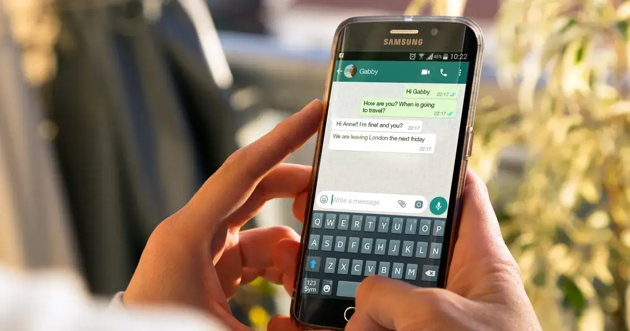 Dłonie trzymają smartfon z otwartym WhatsAppem. Widoczna jest klawiatura i okno czatu, co może pomóc w zrozumieniu, jak zainstalować WhatsApp.