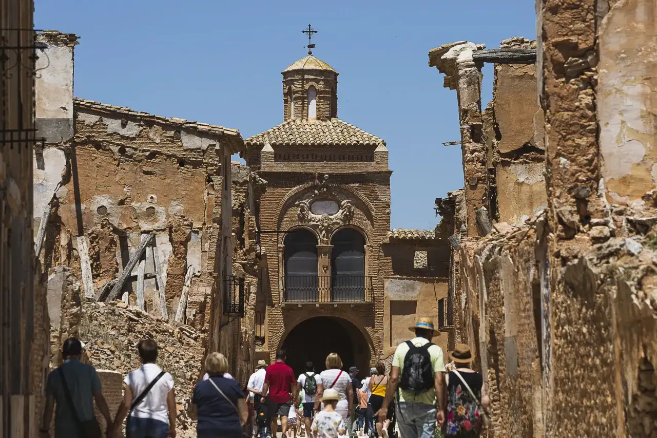 Gente camina por las ruinas de Belchite Viejo, un pueblo fantasma con edificios derruidos y una iglesia en el centro.