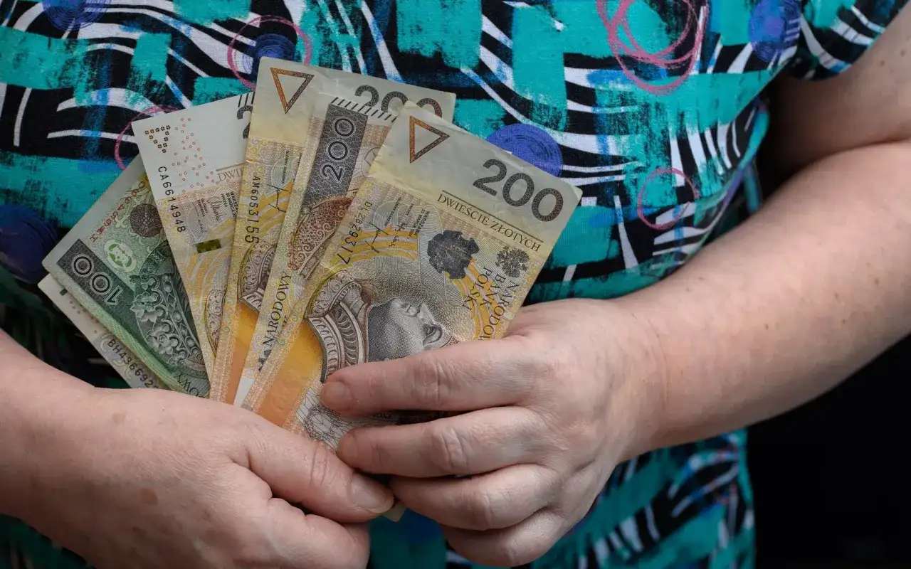 Dłonie trzymają polskie banknoty 100 i 200 zł. Może to być pomoc finansowa, np. specjalny zasiłek opiekuńczy.