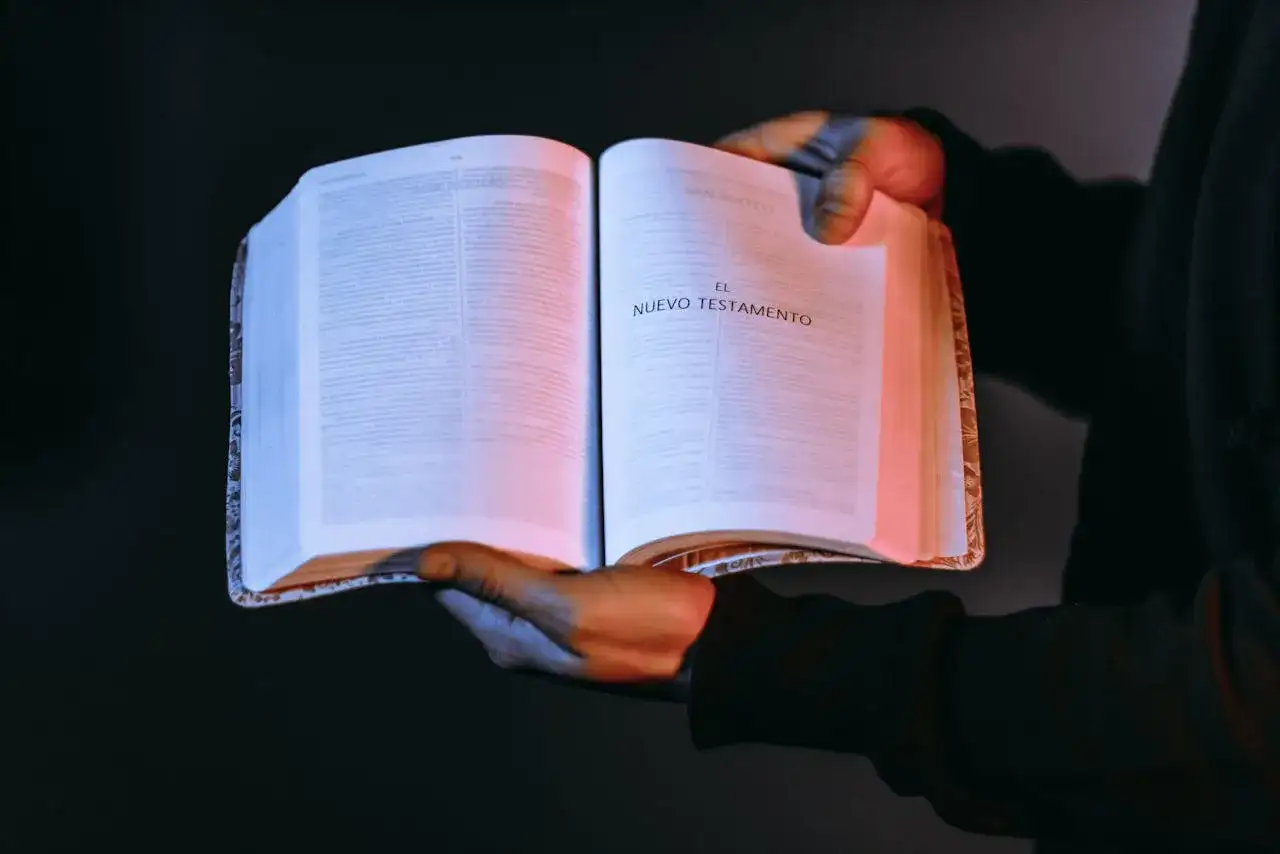 Otwarta księga z napisem "El Nuevo Testamento". W rękach trzymających ją można rozpoznać autor dziejów apostolskich.