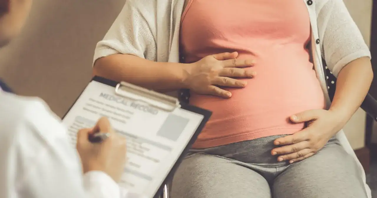 Lekarz notuje dane pacjentki, która trzyma dłoń na brzuchu. Obliczenia terminu porodu opierają się na regule Naegelego.