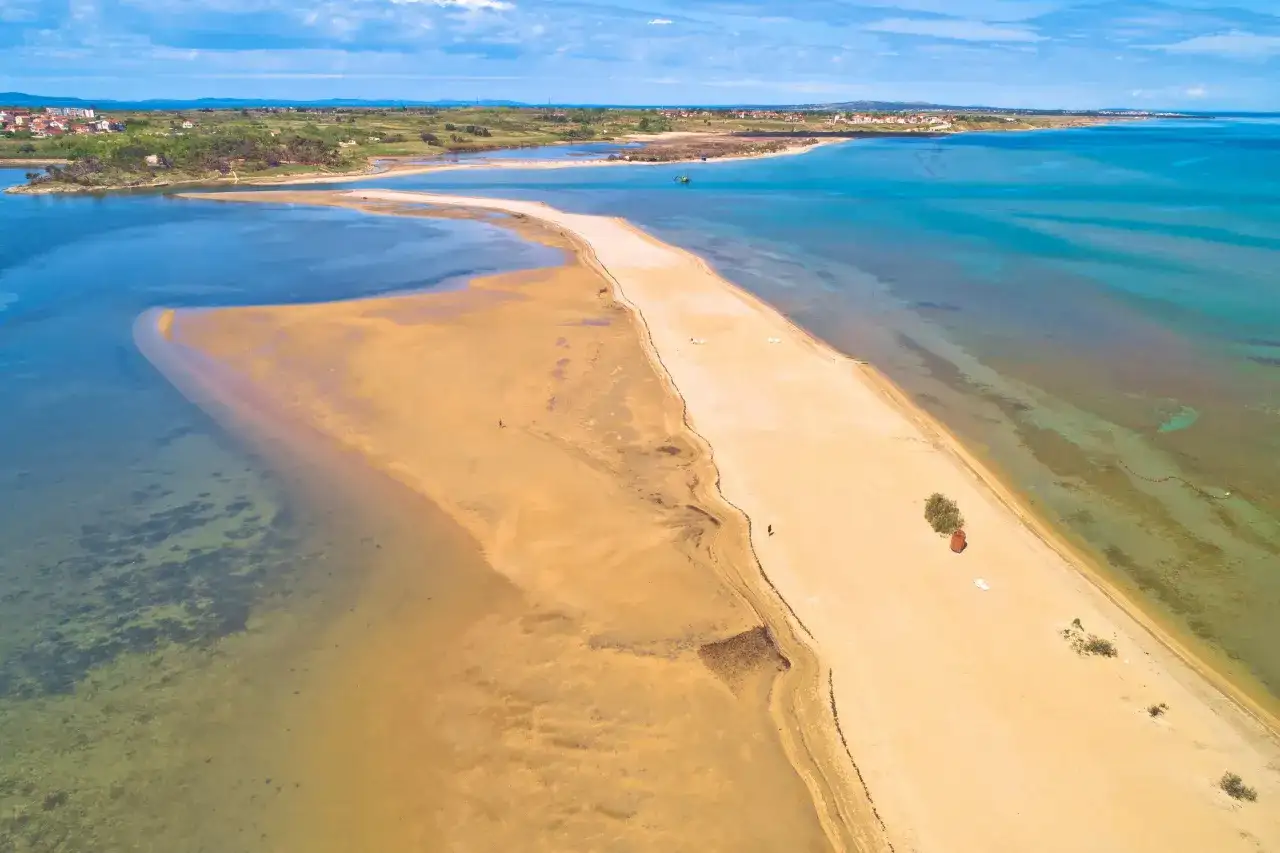 Piaszczyste plaże Chorwacji w całej okazałości. Długi, piaszczysty cypel otoczony turkusową wodą, z widokiem na miasteczko w oddali.