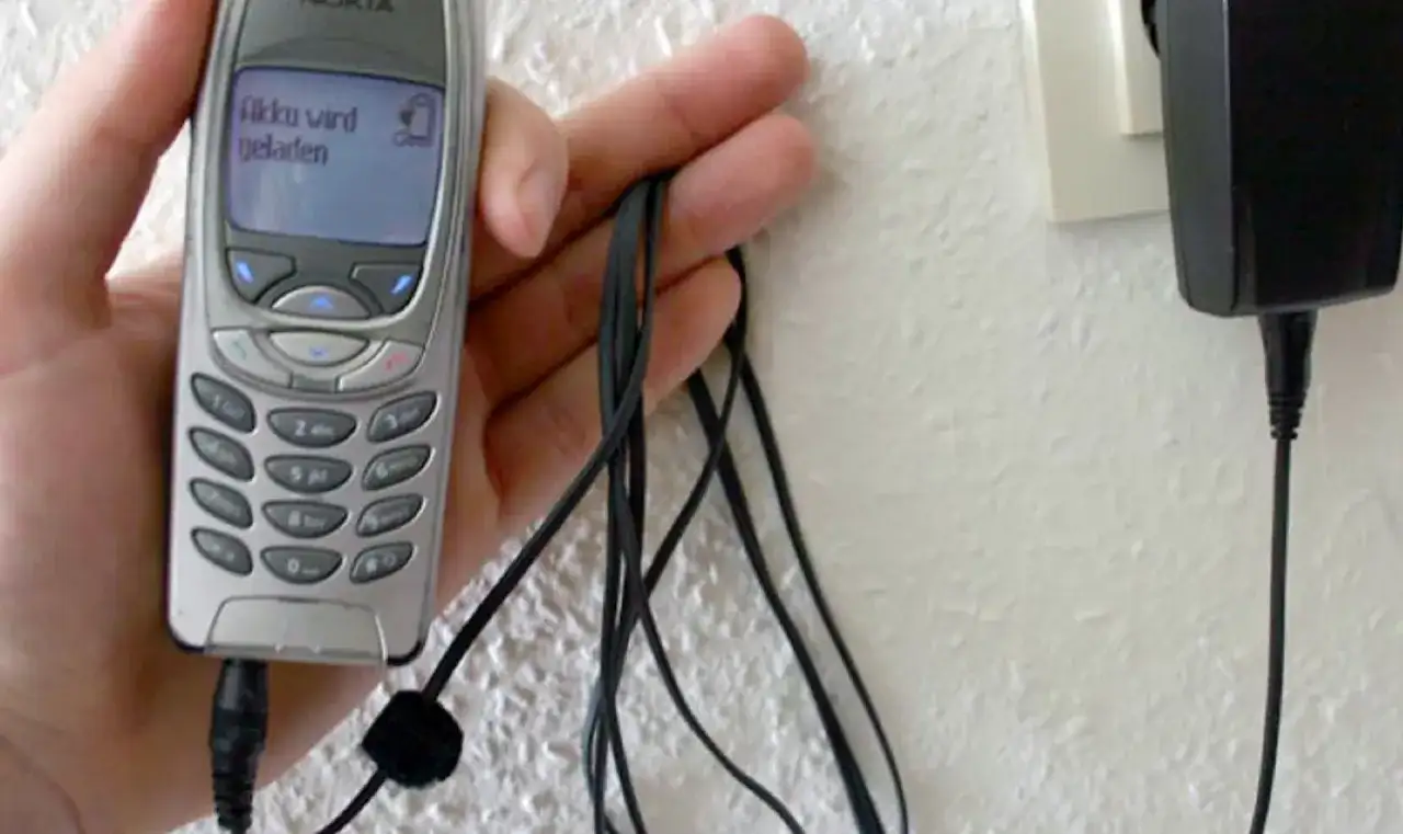Dłoń trzyma starą Nokię, która ładuje się przez kabel. Na ekranie telefonu widnieje napis "Akku wird geladen". To klasyczny przykład technologii 3G z dawnych lat.