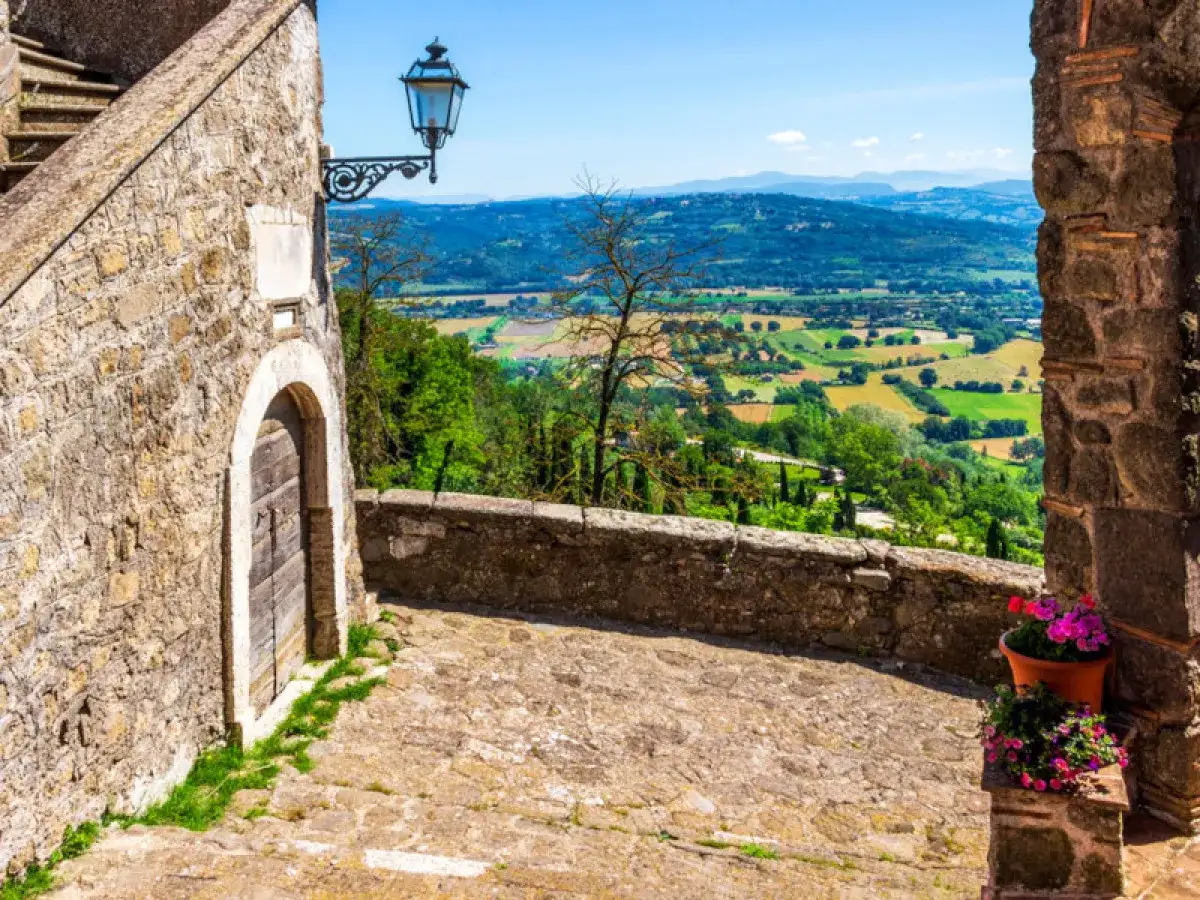 Un vicolo acciottolato a Bassano in Teverina conduce a un panorama mozzafiato di colline verdi e campi coltivati sotto un cielo azzurro.