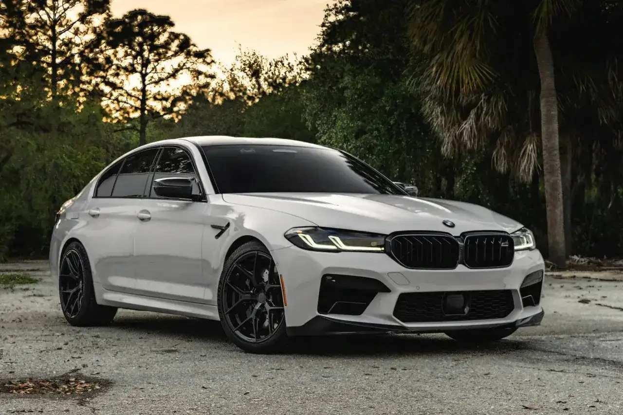 Białe BMW M5 G30 z czarnymi felgami i detalami, zaparkowane na tle drzew i zachodzącego słońca.