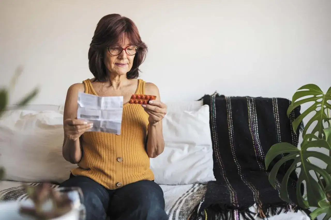 Kobieta czyta ulotkę leku, trzymając opakowanie tabletek. Szuka informacji o lekach na menopauzę bez recepty.