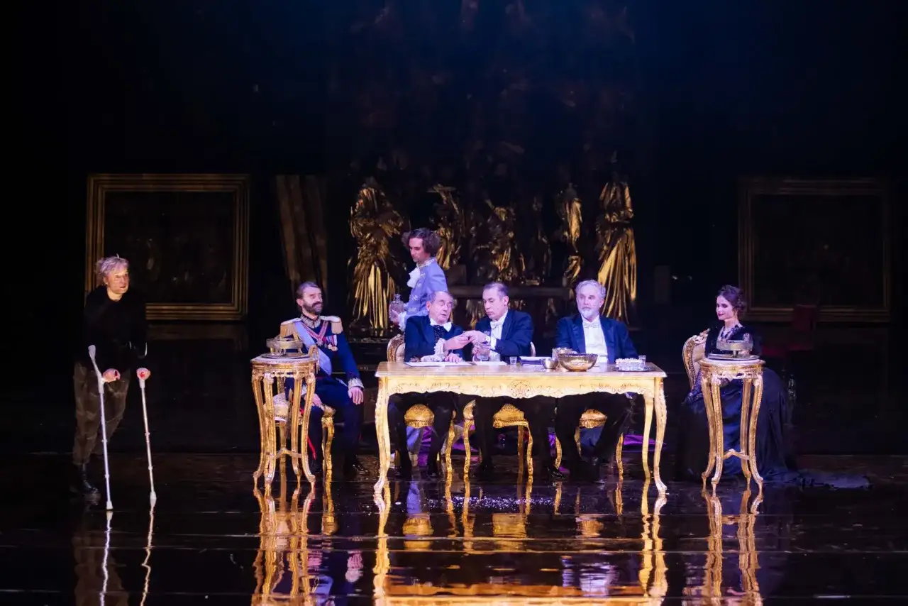 Scena z "Dziadów" w warszawskim teatrze. Przy złotym stole siedzą mężczyźni w strojach z epoki, obok kobieta na wózku.