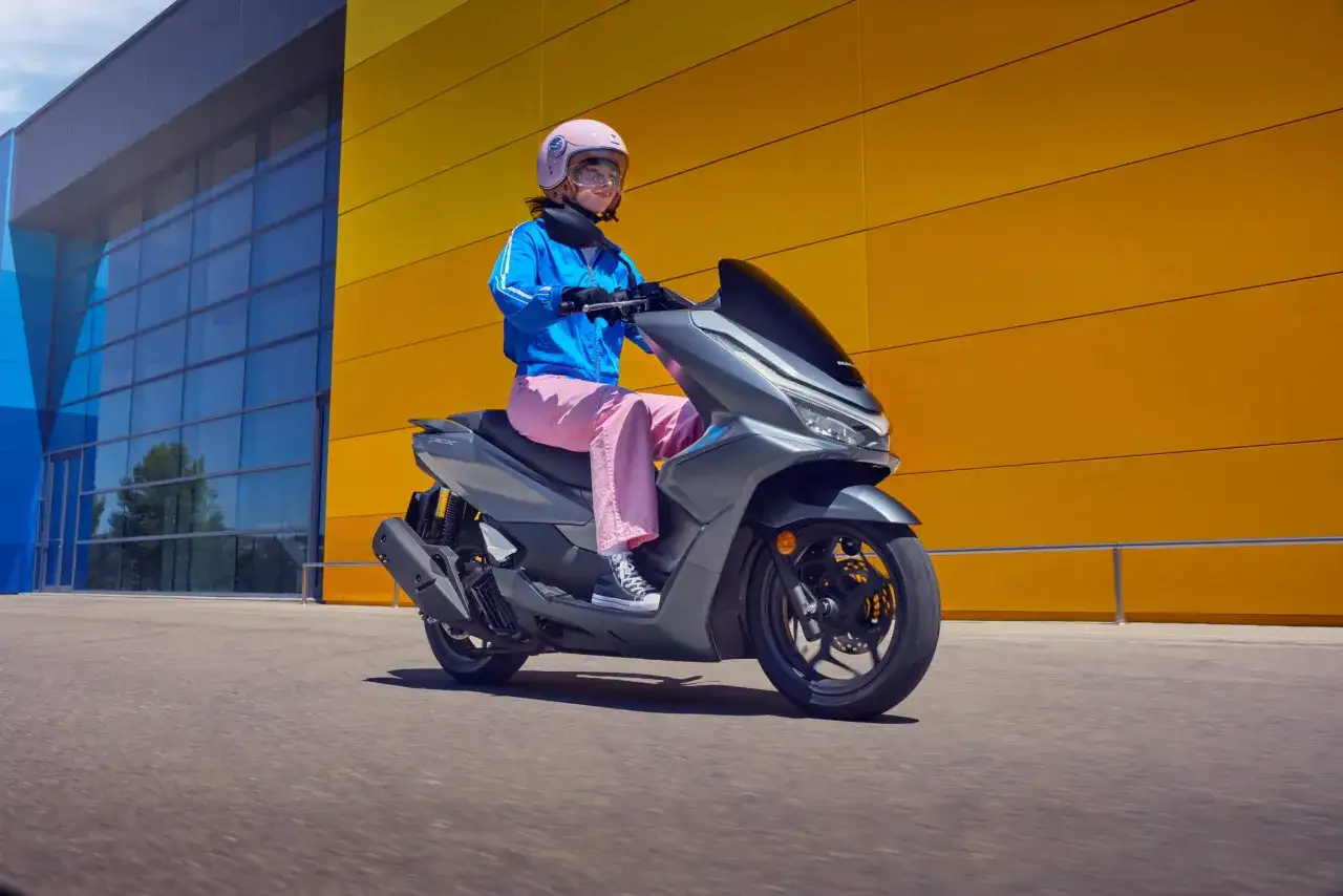 Kobieta w różowych spodniach i niebieskiej kurtce jedzie na szarym skuterze Honda PCX 125. Wady tego modelu mogą dotyczyć np. zawieszenia.