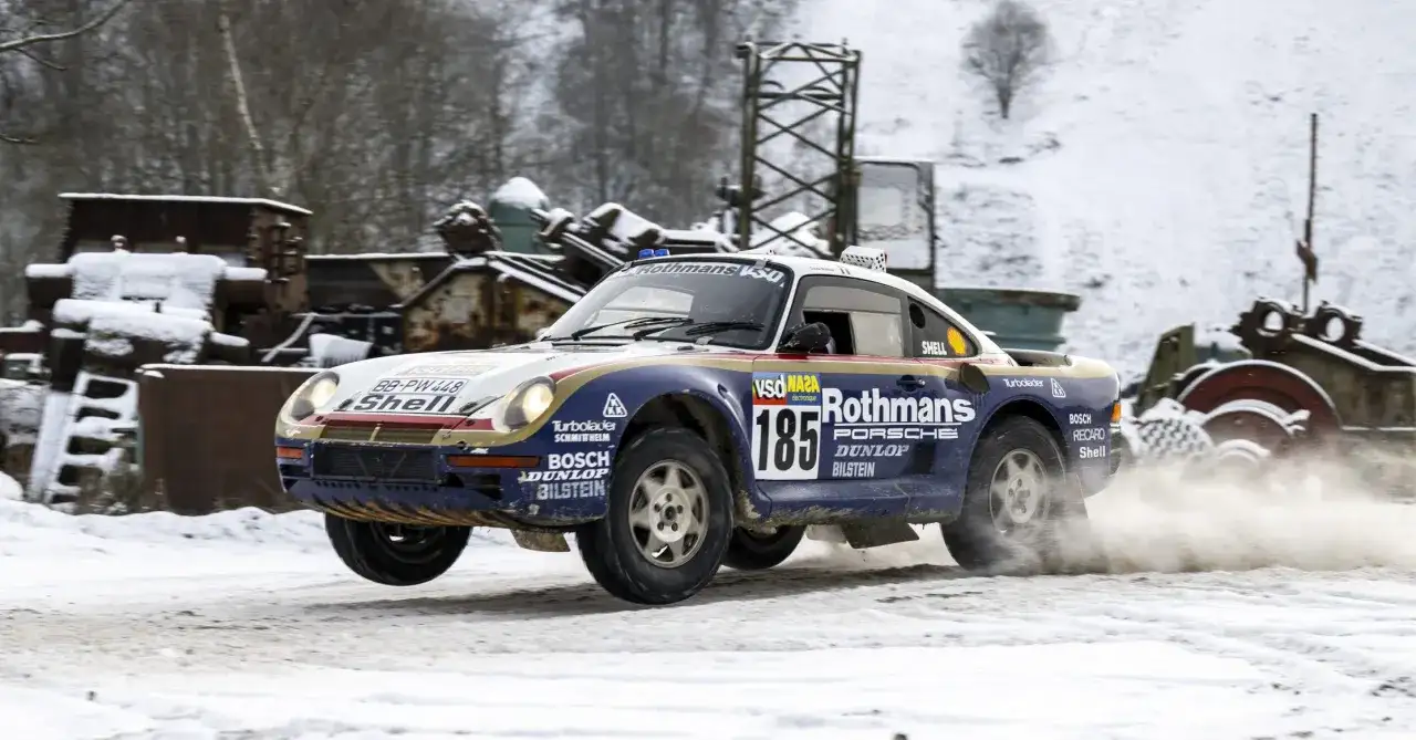 Porsche 959 w barwach Rothmans, z numerem 185, pokonuje śnieżny odcinek rajdu, wzbijając tumany śniegu.