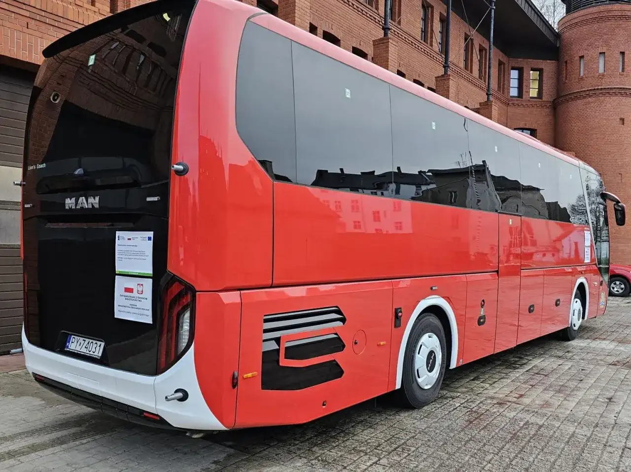 Czerwony autokar MAN Lion's Coach zaparkowany przed ceglanym budynkiem. Może to być przystanek w podróży z Bolesławca do Poznania.