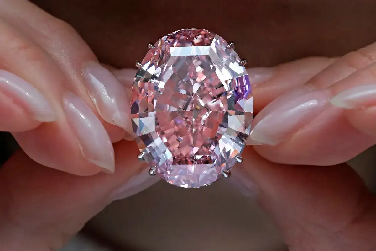 Dłonie trzymają olśniewający, owalny, różowy diament, prawdopodobnie najdroższy diament na świecie, błyszczący w świetle.