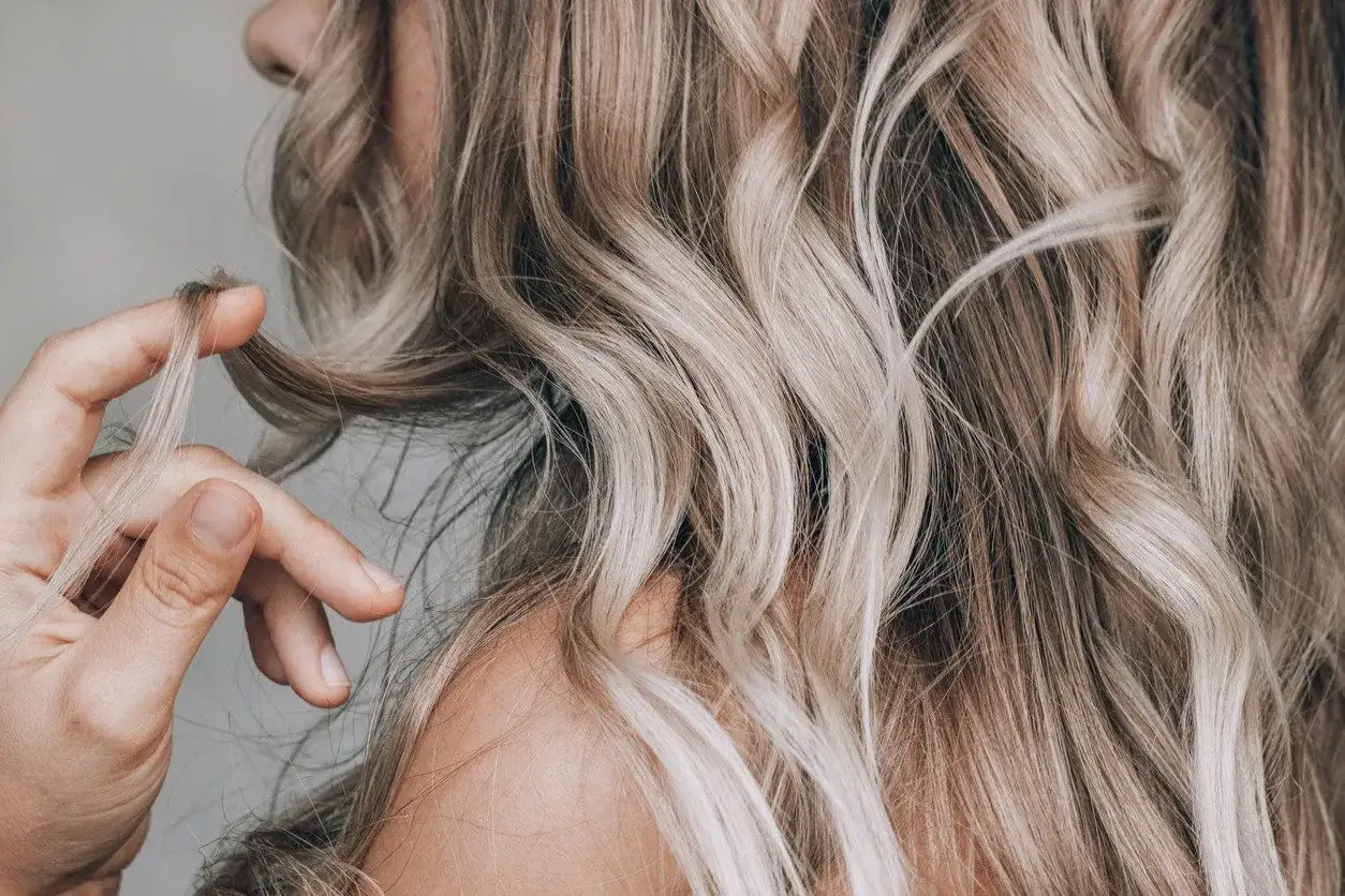 Dłoń dotyka falujących włosów w odcieniu sombre blond, podkreślając ich naturalne przejścia kolorystyczne.
