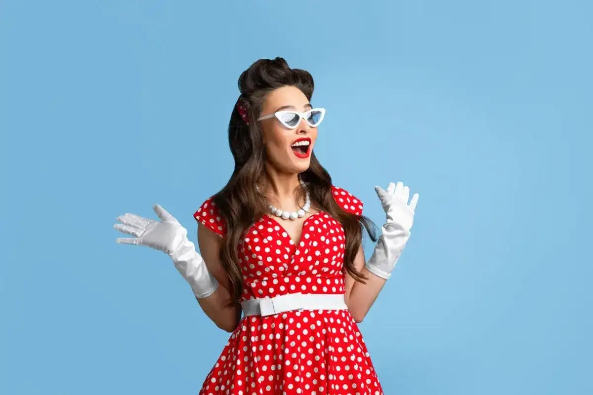 Kobieta w stylu pin-up w czerwonej sukience w białe grochy, białych rękawiczkach i okularach, z uniesionymi rękami.