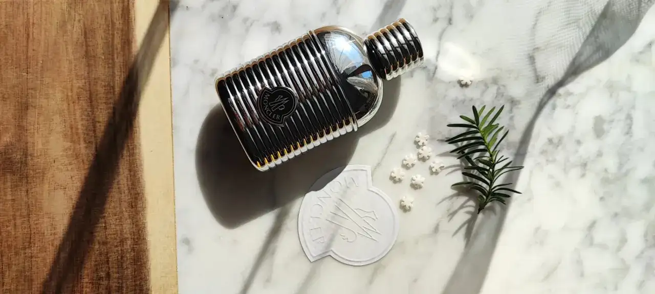 Elegancki flakon perfum Moncler pour homme na marmurowym tle, otoczony gałązką iglaków i płatkami śniegu.