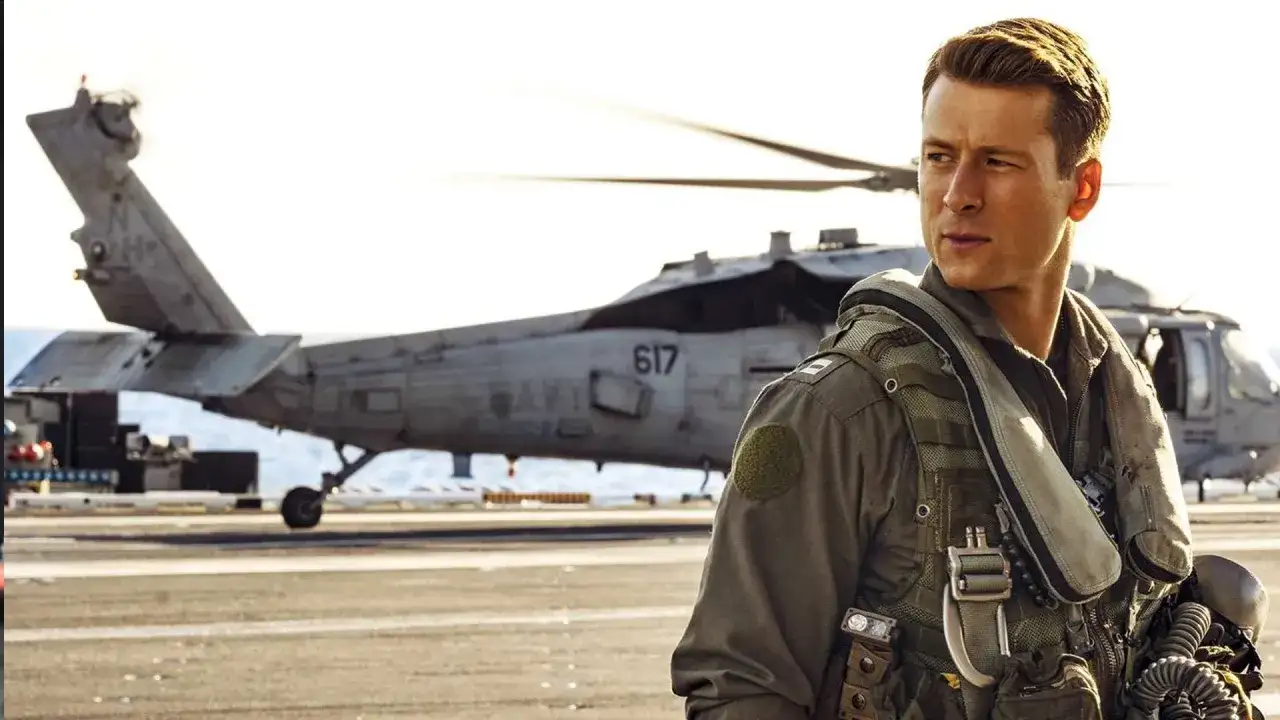 Glen Powell w filmach jako pilot na pokładzie lotniskowca, z helikopterem w tle.