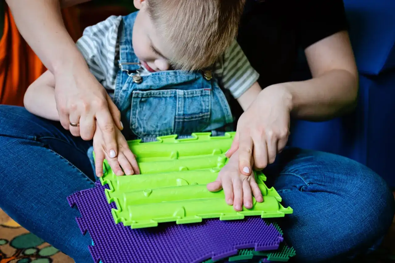 Dziecko w ogrodniczkach z pomocą terapeuty bawi się sensorycznymi matami. Terapia dla dzieci z problemami emocjonalnymi często wykorzystuje takie zabawy.