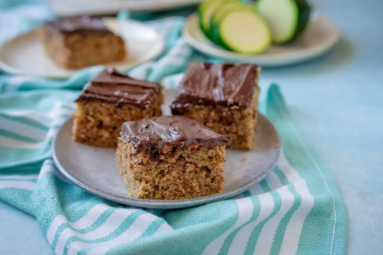 Saftiger Zucchini Kuchen mit Schokoladenglasur auf einem Teller, daneben ein paar Zucchinischeiben.