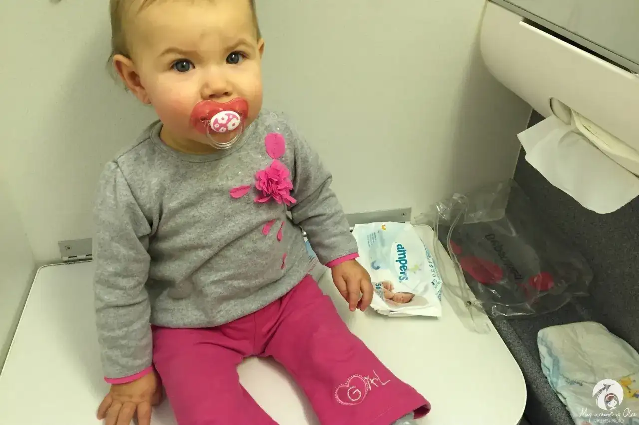 Mała dziewczynka z smoczkiem siedzi na siedzeniu w samolocie Ryanair. Obok niej leżą pieluchy i chusteczki.