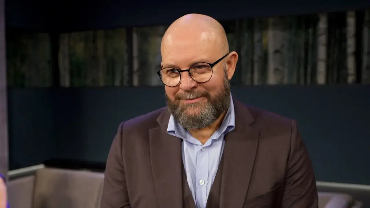 Szymon Bobrowski w garniturze i okularach, z brodą, wygląda jakby opowiadał o swoich filmach, serialach i programach.