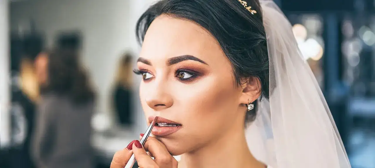 Panna młoda z welonem i biżuterią otrzymuje makijaż ślubny glamour. Usta podkreślone błyszczykiem, oczy smoky eye.