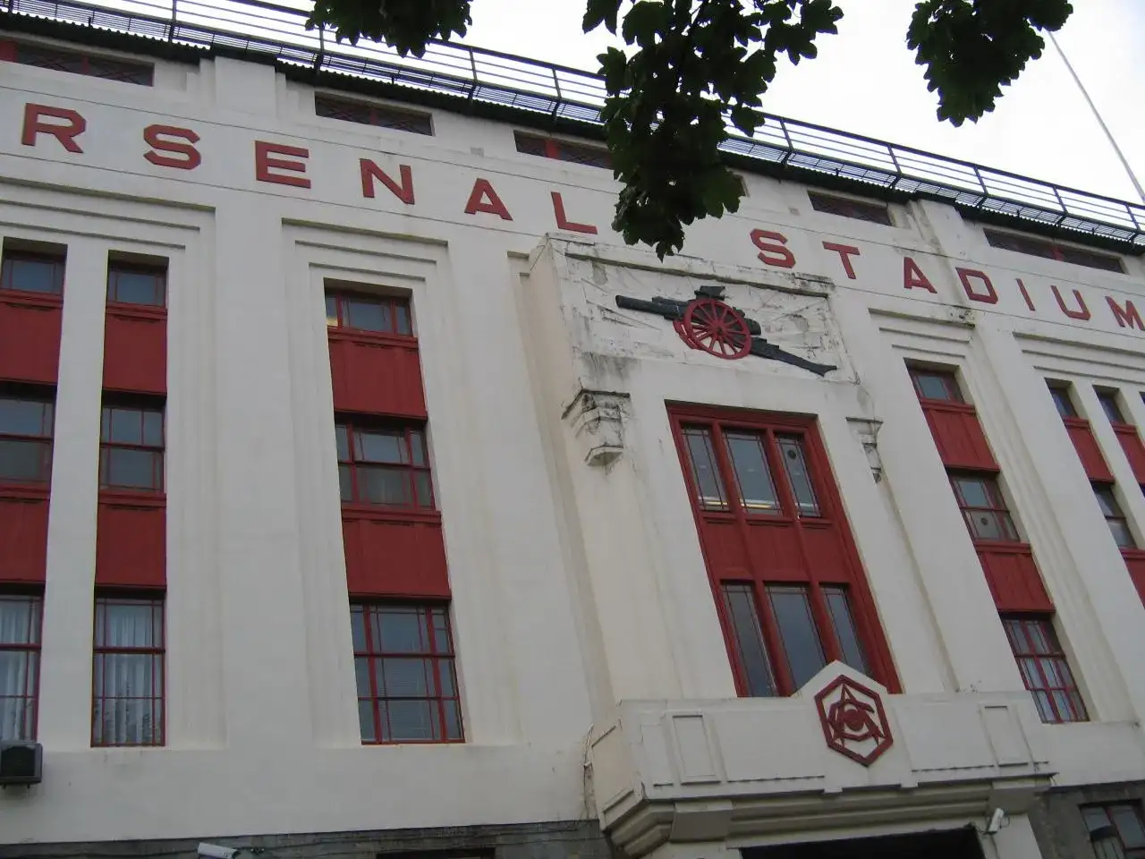 Stary stadion Arsenalu z czerwonymi oknami i napisem "ARSENAL STADIUM". Widoczny herb klubu i armata.