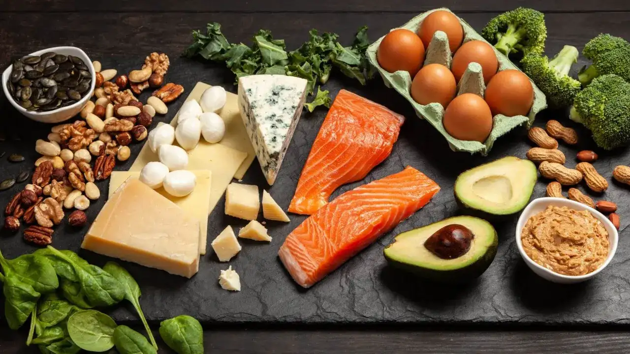 Na talerzu składniki na dietę keto: łosoś, awokado, jajka, sery, orzechy, brokuły i szpinak.