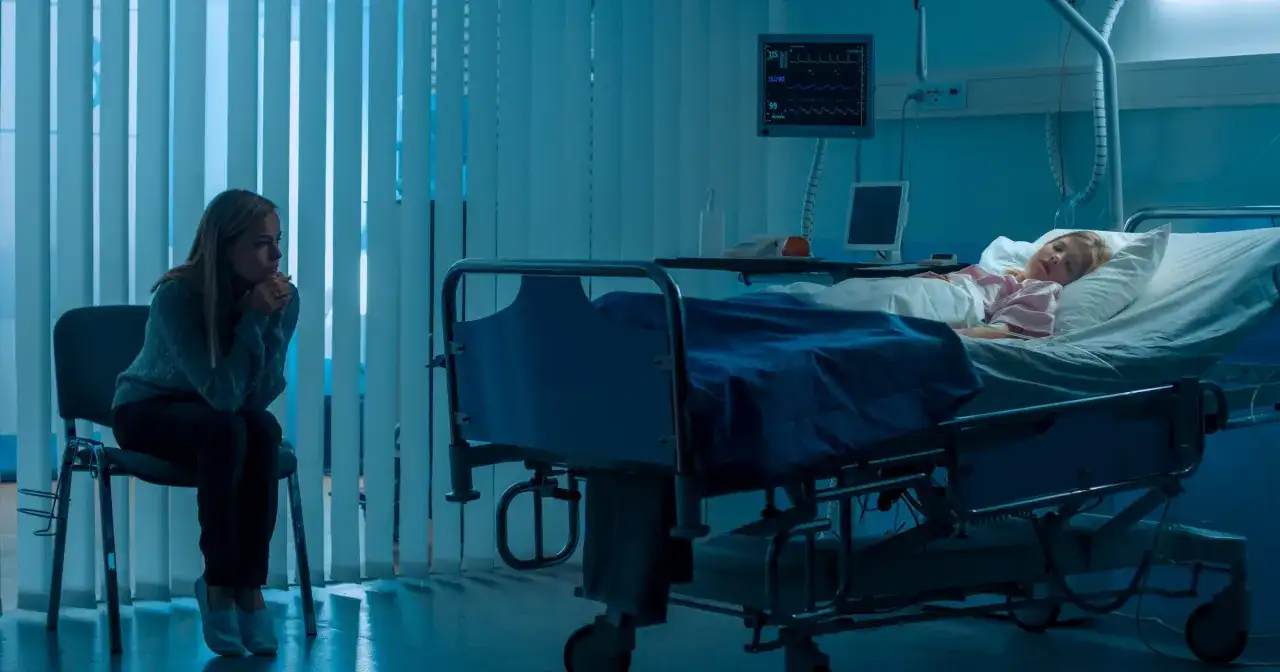Zmartwiona matka siedzi obok śpiącego dziecka w szpitalnym łóżku, co może sugerować zespół Münchhausena u matek.