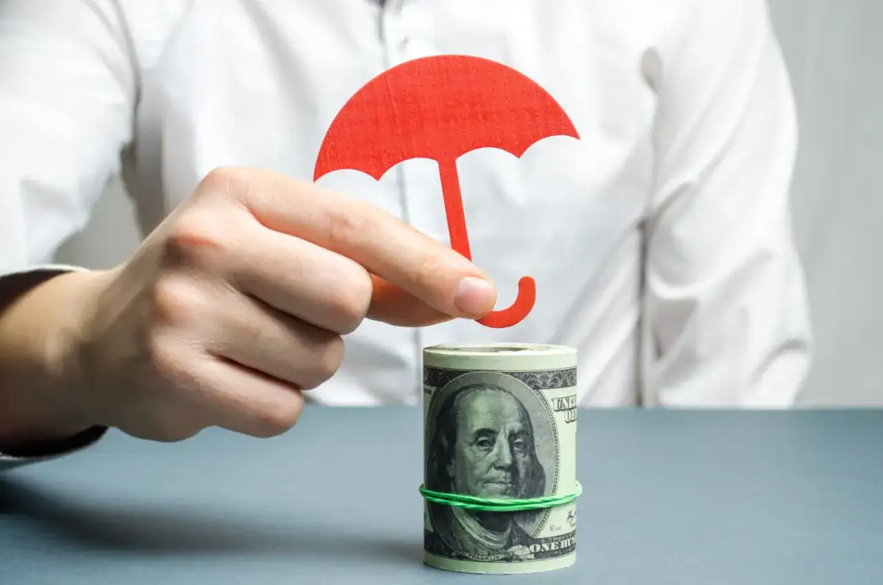 Dłoń trzyma czerwony parasol nad zrolowanymi dolarami. Tani murek oporowy dla Twoich finansów.