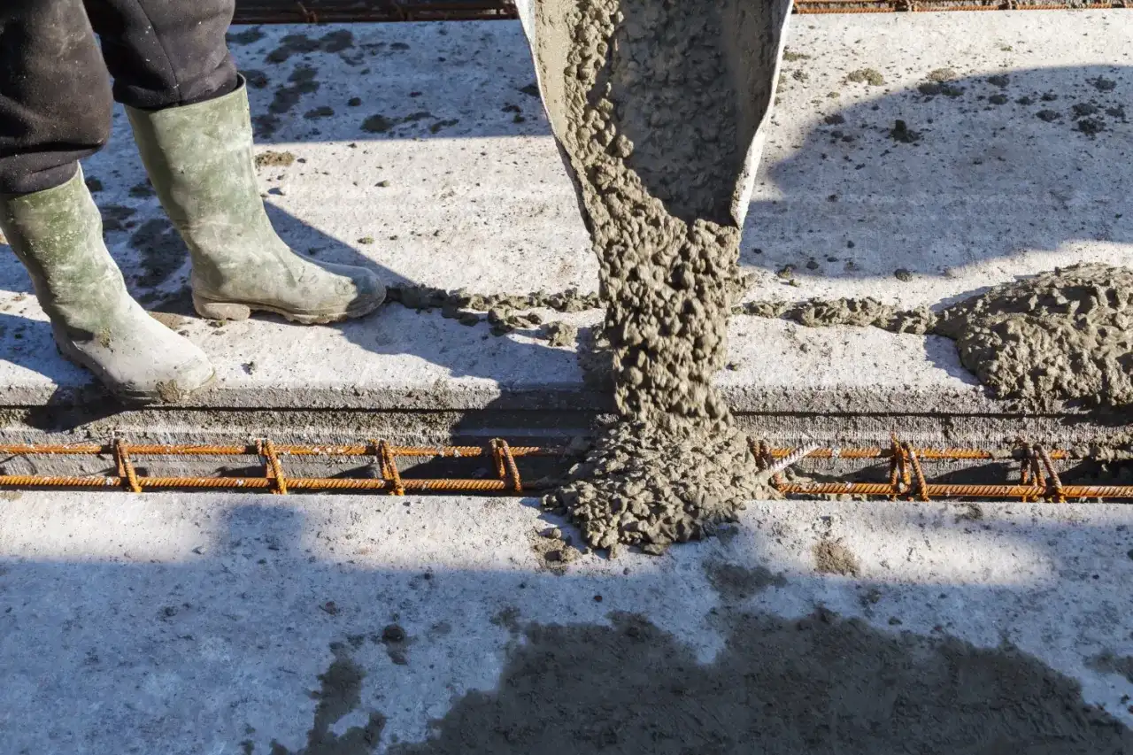 Beton wylewany z rury na zbrojenie. Pracownik w gumowcach stoi obok, nadzorując proces.