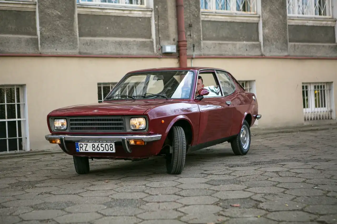 Czerwony fiat 128, klasyk z lat 70., zaparkowany na brukowanym dziedzińcu przed budynkiem.