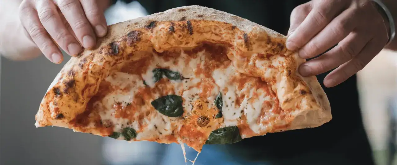 Hände halten ein Stück Pizza mit luftigem Rand, geschmolzenem Käse und Basilikum.