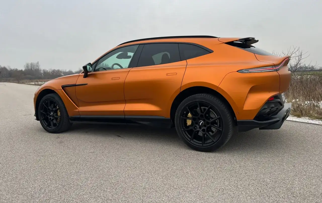 Pomarańczowy Aston Martin DBX z czarnymi felgami i przyciemnianymi szybami na tle pochmurnego nieba.