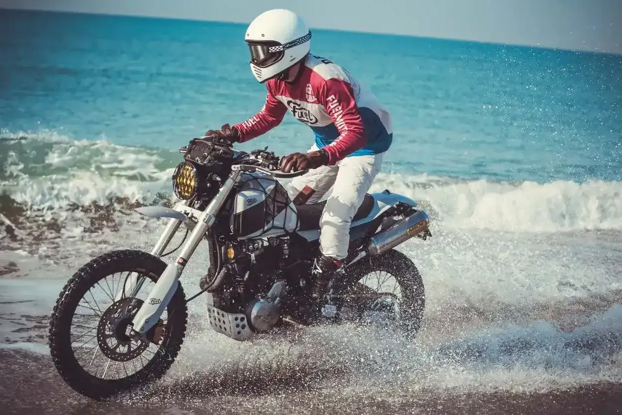 Motocyklista w kasku i kolorowej bluzie śmiga na Honda FMX 650 po plaży, wzbijając fontanny wody.