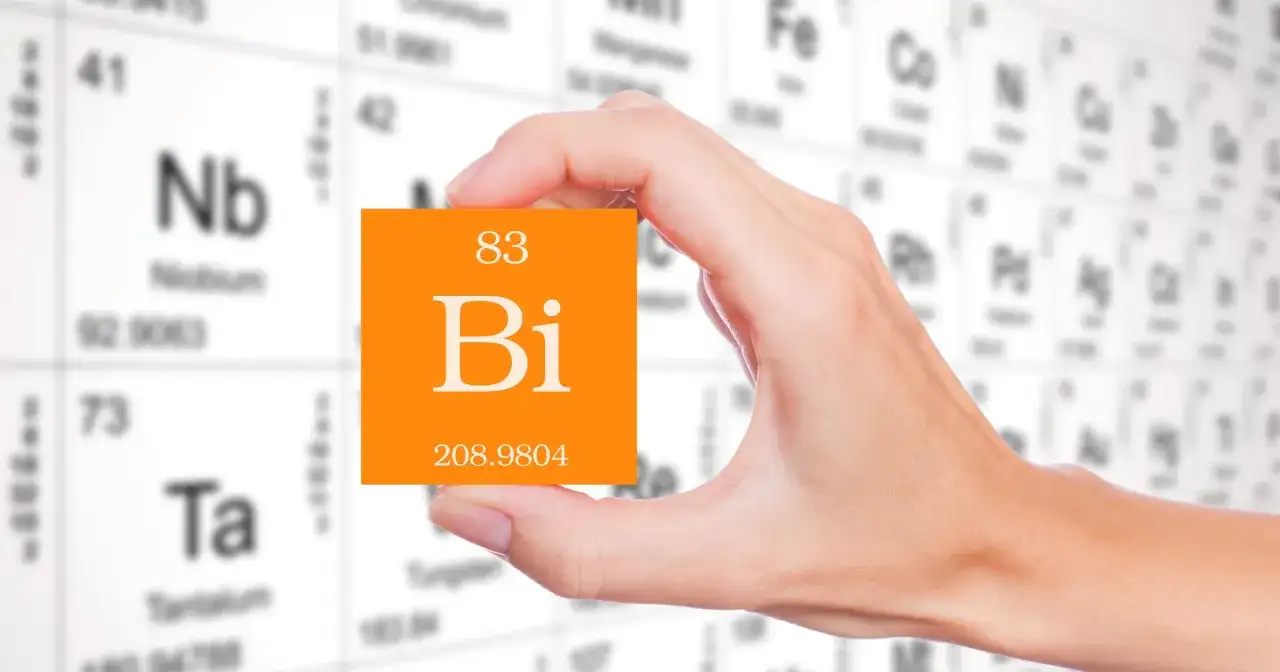 Dłoń trzyma pomarańczową kartkę z symbolem pierwiastka bizmut (Bi) i jego liczbą atomową 83. W tle widać tablicę Mendelejewa.