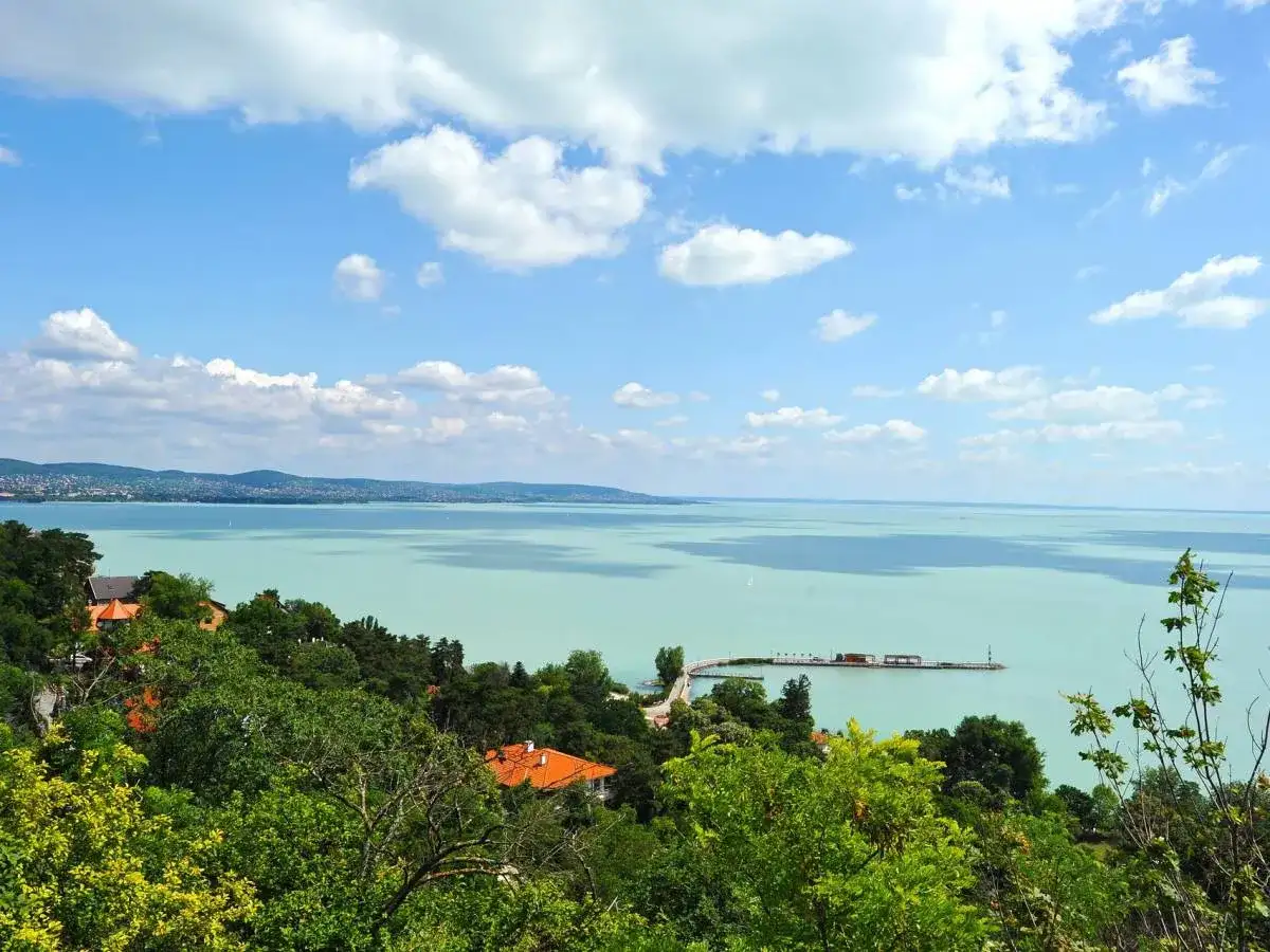 Widok na turkusowe wody Balatonu z zielonymi wzgórzami i domami. To jedno z najpiękniejszych miejsc nad Balatonem, z molo i żaglówkami na horyzoncie.