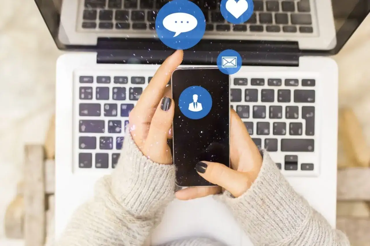Dłonie trzymające smartfon nad klawiaturą laptopa. Ikony komunikatorów unoszą się nad ekranem. Chcesz wiedzieć, jak sprawdzić kiedy ktoś był aktywny na Messengerze?
