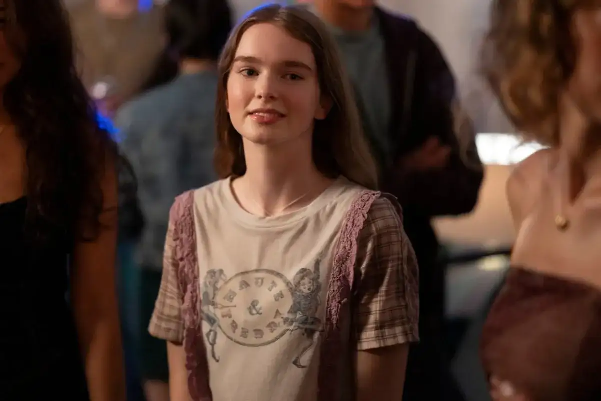Młoda dziewczyna w koszulce z nadrukiem, przypominająca postać z filmów Chloë Grace Moretz, uśmiecha się na imprezie.