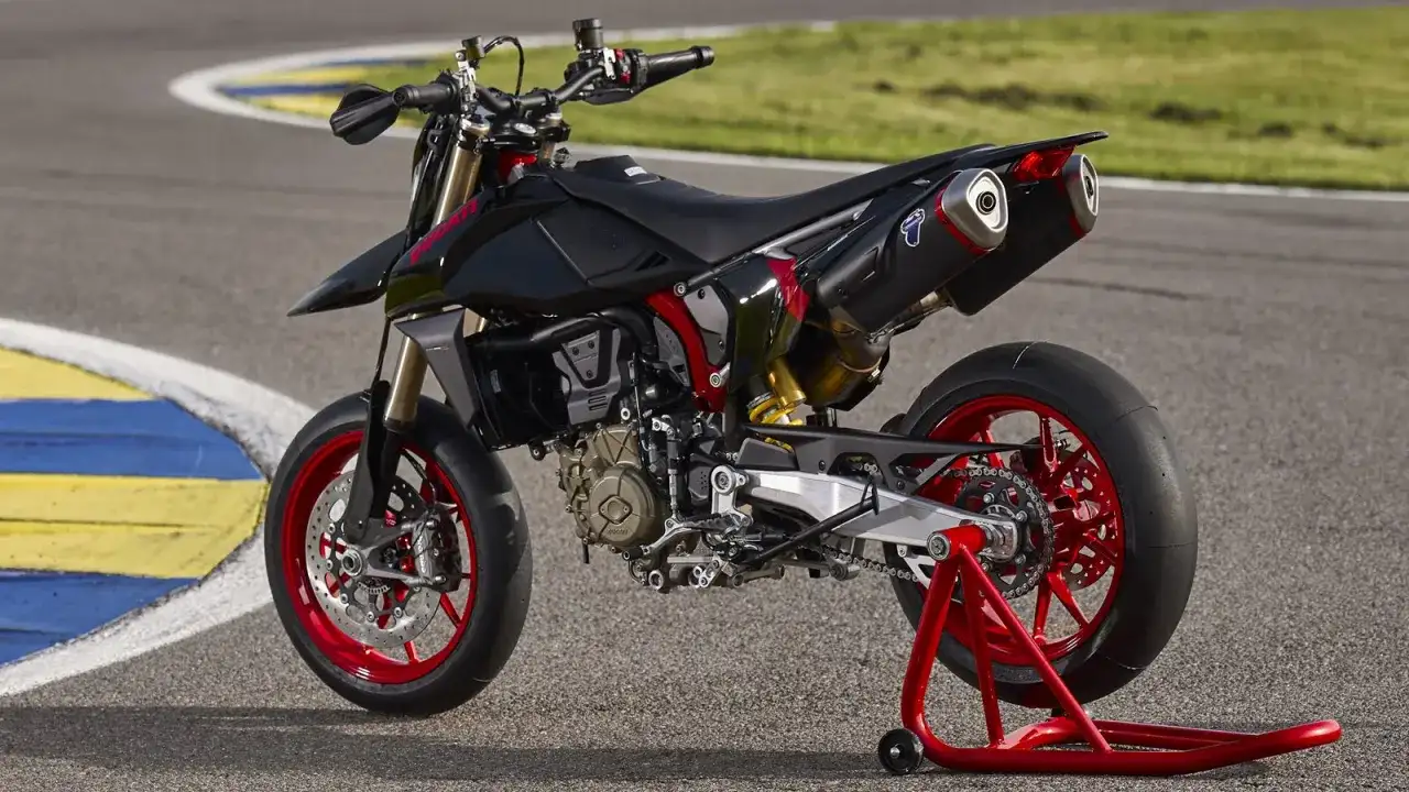 Czarny motocykl **hypermotard** z czerwonymi felgami i akcentami, stojący na czerwonym stojaku na torze wyścigowym.