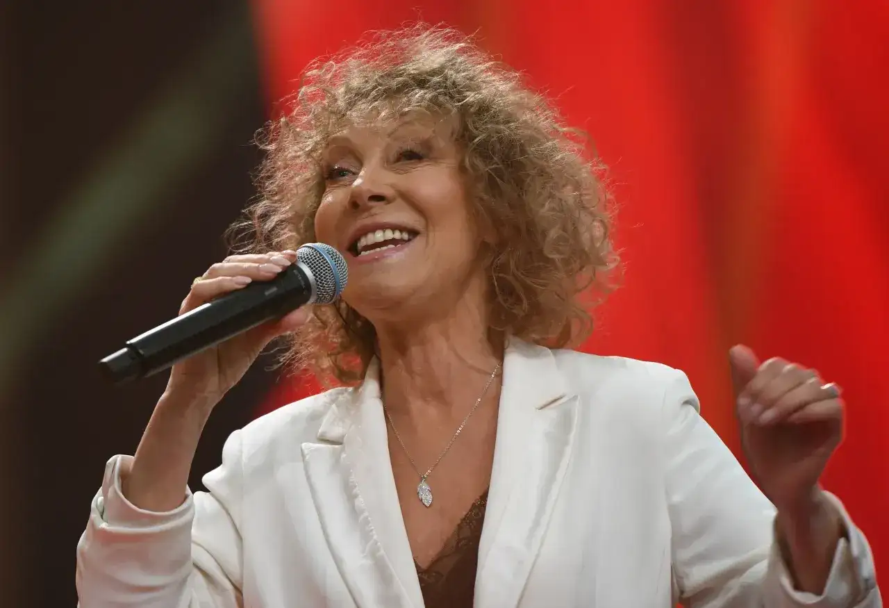 Alicja Majewska, mimo swojego wieku, wciąż zachwyca na scenie. Uśmiechnięta, z mikrofonem w dłoni, w białej marynarce.