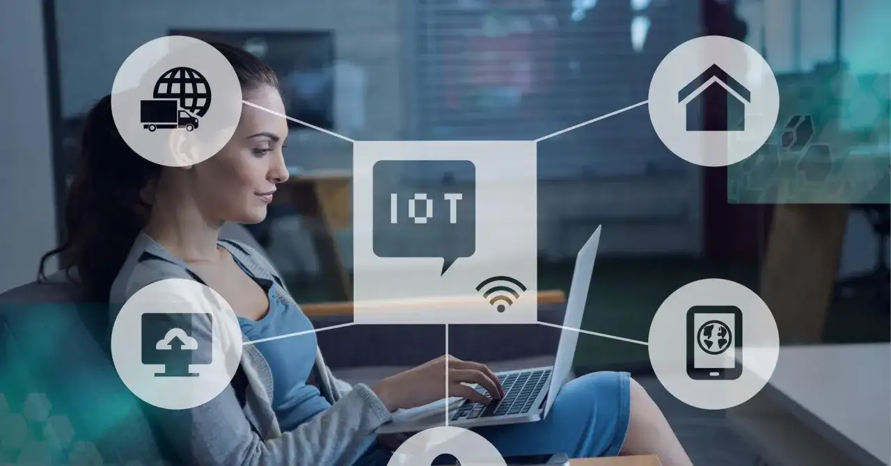 Kobieta pracuje przy laptopie, otoczona ikonami symbolizującymi internet rzeczy (IoT): dom, chmura, ciężarówka, smartfon.