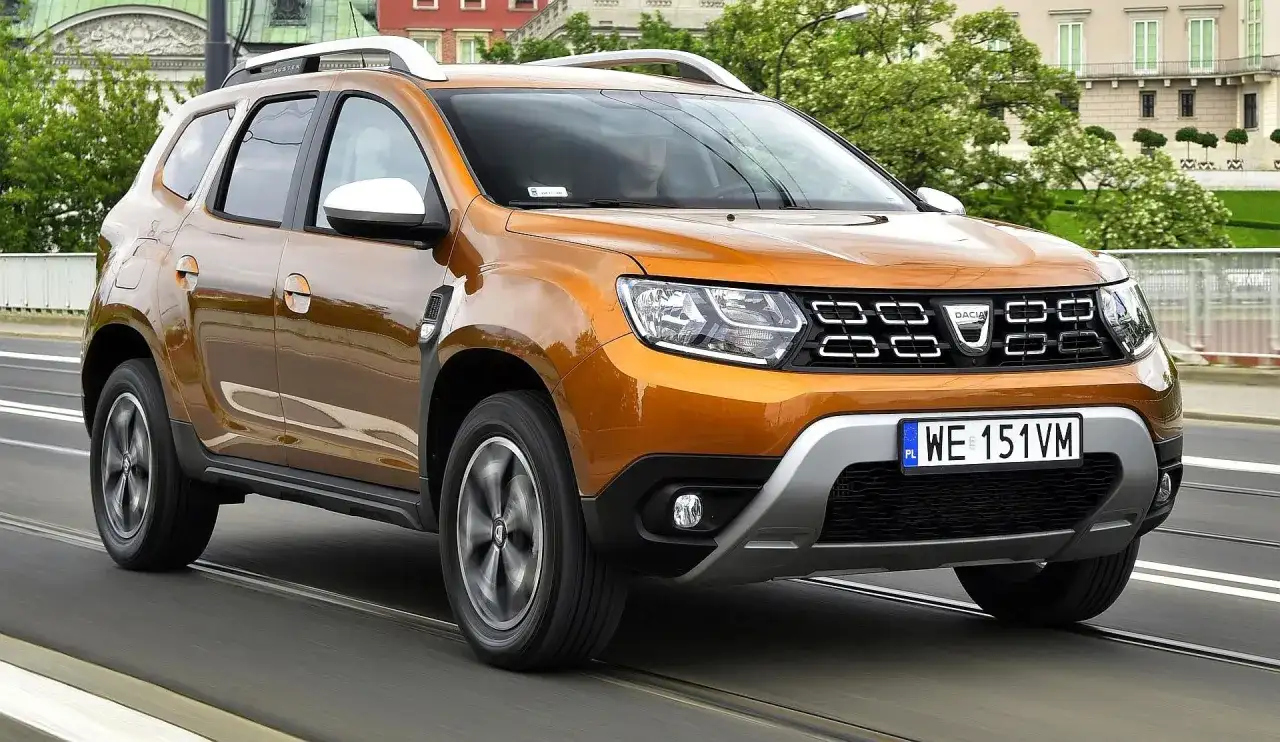 Pomarańczowy Dacia Duster 2 jedzie po moście, za nim widać budynki i drzewa.