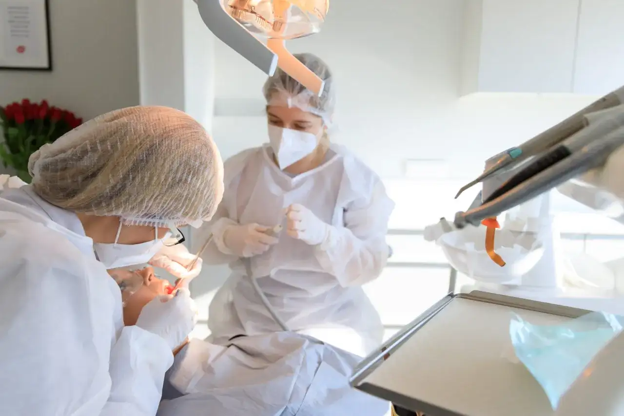 Dentystka w masce i fartuchu wykonuje zabieg kiretażu u pacjentki. W tle widać sprzęt stomatologiczny.