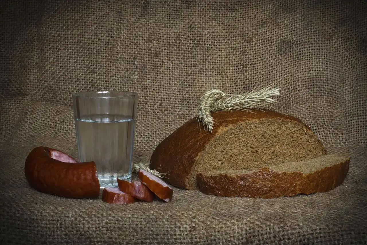Szklanka wódki z ziemniaka, kromka chleba i kiełbasa na płóciennym tle. Proste, polskie smaki.